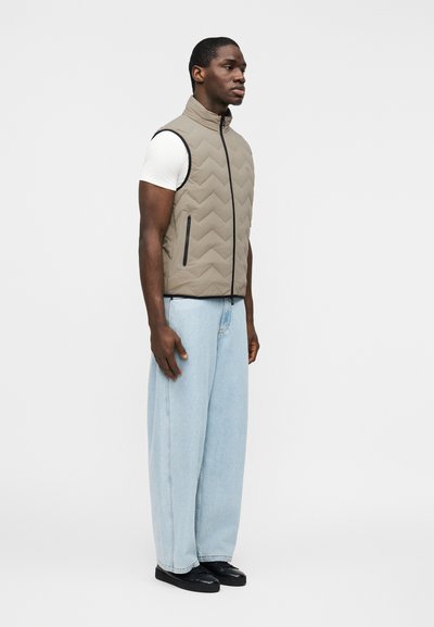 Άνδρας με μπεζ γιλέκο με ραφές πάνω από λευκό κοντομάνικο πουκάμισο, φαρδιά ανοιχτό μπλε τζιν και μαύρα παπούτσια, στέκεται μπροστά σε λευκό απλό φόντο.