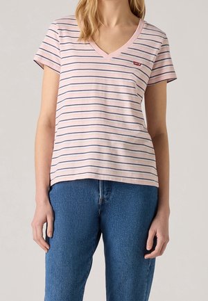 Femme portant un t-shirt rose à col en V avec de fines rayures bleu marine et un jean bleu, debout devant un fond clair uni.
