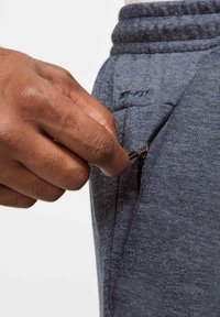 Pantalones deportivos grises con una superficie texturizada, que cuentan con un bolsillo con cremallera y una cinturilla elástica. Mano extendiéndose hacia la cremallera.