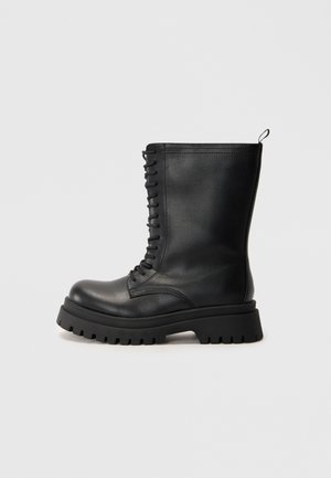 Stivale in pelle nera con lacci, suola in gomma spessa, punta rotonda e una linguetta sul retro. Presenta cuciture verticali lungo i lati.