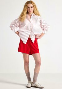Röda shorts med dragsko i midjan, tillsammans med en vit knapp-up skjorta med röda vertikala ränder. Krämfärgade sneakers och taupe-strumpor fulländar looken.