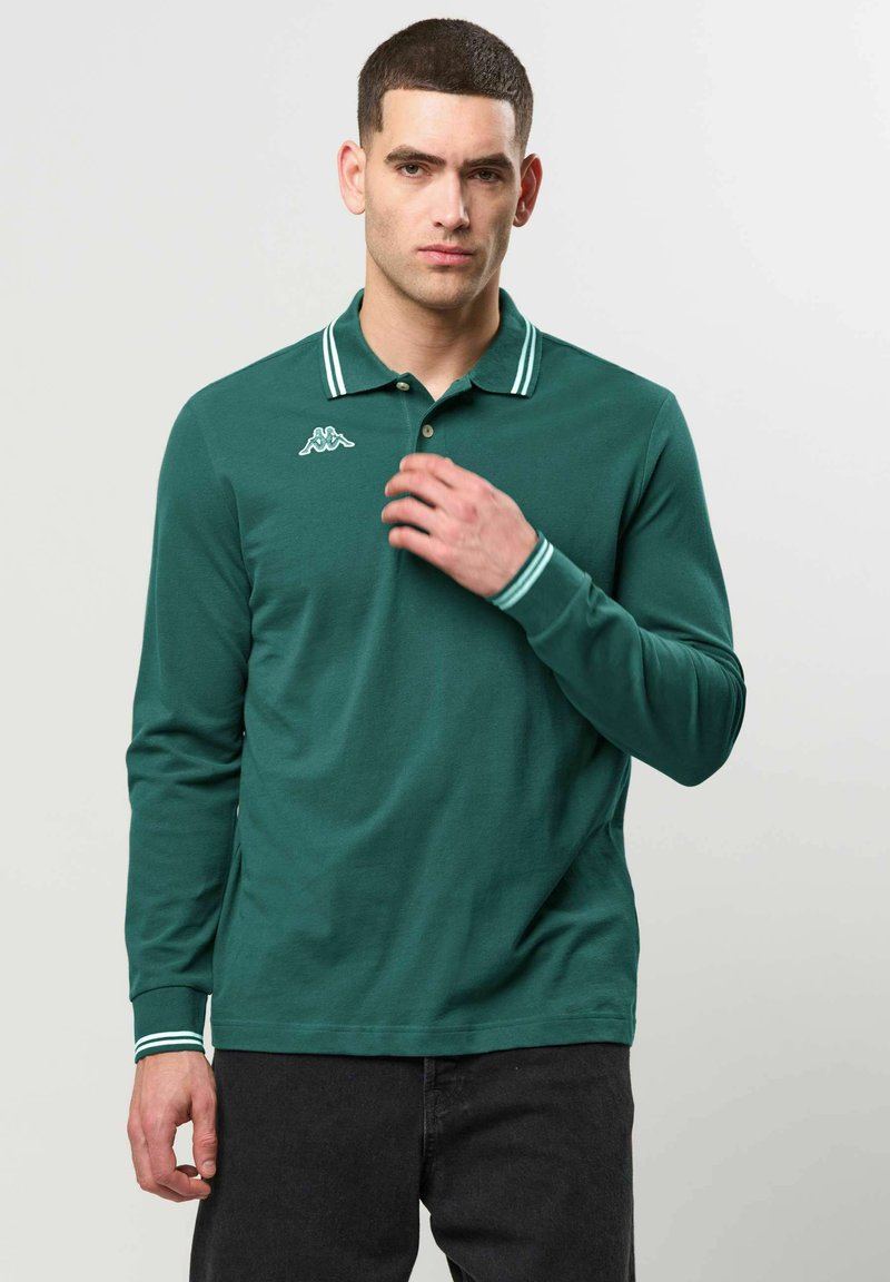Homme aux cheveux courts et foncés, portant un polo vert à manches longues avec des rayures blanches sur le col et les poignets, et un pantalon noir, debout sur un fond uni.