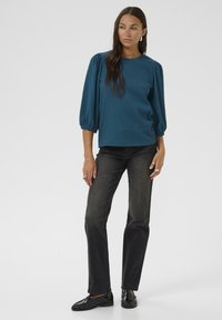 Blouse turquoise texturée avec des manches bouffantes, associée à un jean droit noir et des mocassins noirs. Le modèle se tient contre un fond blanc.
