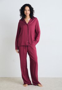 Burgunderfärgad pyjamasuppsättning med vertikala ränder, som har en knapp-up topp och byxor med dragsko, tillverkad av ett mjukt tyg med en avslappnad passform.