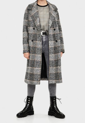 Osoba v dlouhém černobílém kostkovaném kabátu, šedých džínech s vysokým pasem, černém pásku a černých šněrovacích botách stojící na bílém pozadí.