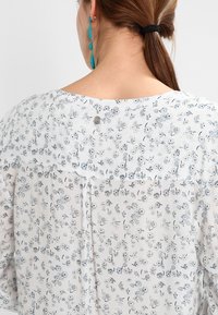 Blouse blanche avec un imprimé floral aux tons bleus et gris, dotée d'une fermeture boutonnée au dos et d'un tissu doux et texturé.