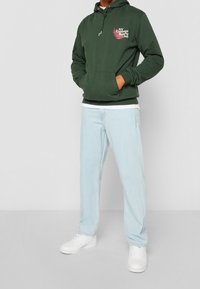 Sudadera verde con capucha, con bolsillo frontal y cordón. Presenta un diseño gráfico en blanco y rosa. Combinada con jeans azul claro y zapatos blancos.