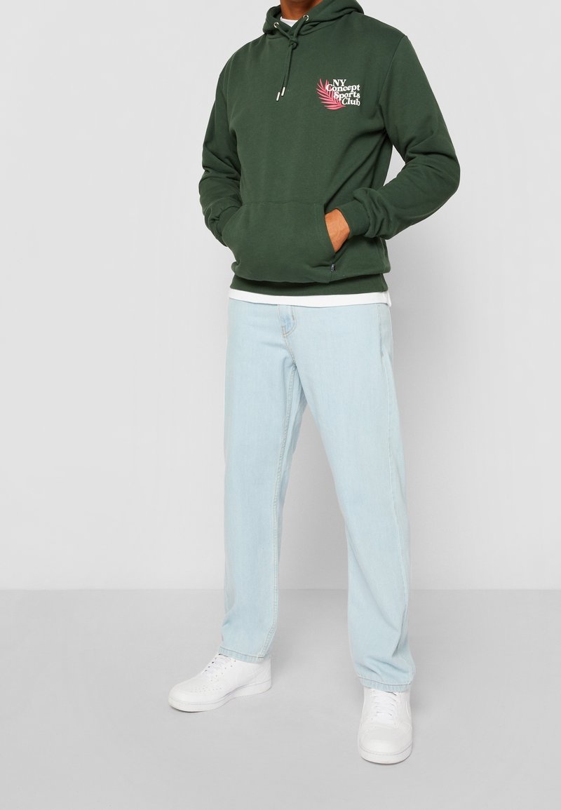 Sudadera verde con capucha, con bolsillo frontal y cordón. Presenta un diseño gráfico en blanco y rosa. Combinada con jeans azul claro y zapatos blancos.