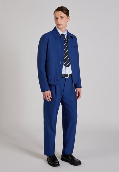 Male model wearing a blue jacket and matching trousers, light blue shirt, striped tie, black belt, and black dress shoes, standing on gray background.