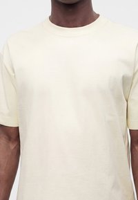 Personne portant un t-shirt uni à manches courtes de couleur crème, visible du milieu de la poitrine au cou sur un fond neutre.