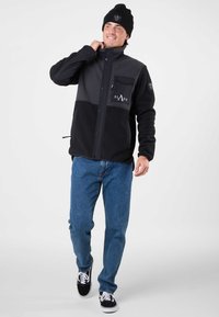 Giacca in pile nera e grigia con chiusura a bottoni, tasca sul petto e dettaglio con logo. Abbinata a jeans blu e sneaker nere.