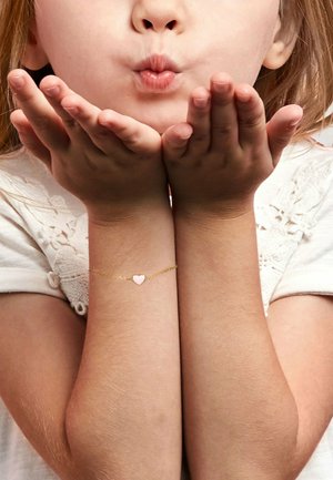 Kind mit heller Haut und rotem Haar, das einen Kuss pustet, trägt ein weißes Hemd und ein goldenes Armband mit einem pinken Herzanhänger am linken Handgelenk.
