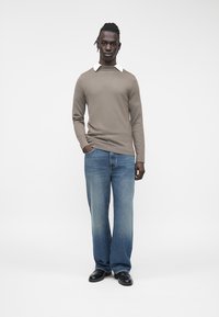 Pull beige à manches longues avec un col blanc, associé à un jean bleu clair à jambes larges et des chaussures noires, debout devant un fond blanc.