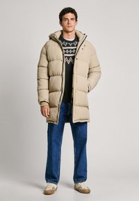 Beige puffer coat with a hood, featuring a front zipper, snap buttons, and ribbed cuffs; worn over a patterned sweater and blue jeans.