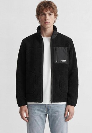 Jeune homme aux cheveux bouclés portant une veste polaire noire zippée par-dessus une chemise blanche et un jean bleu clair, debout devant un fond uni.
