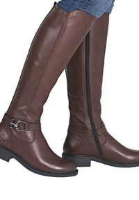 Bottes en cuir marron montant jusqu'au genou avec fermetures éclair latérales noires, sangles décoratives, boucle argentée, portées avec un jean bleu.