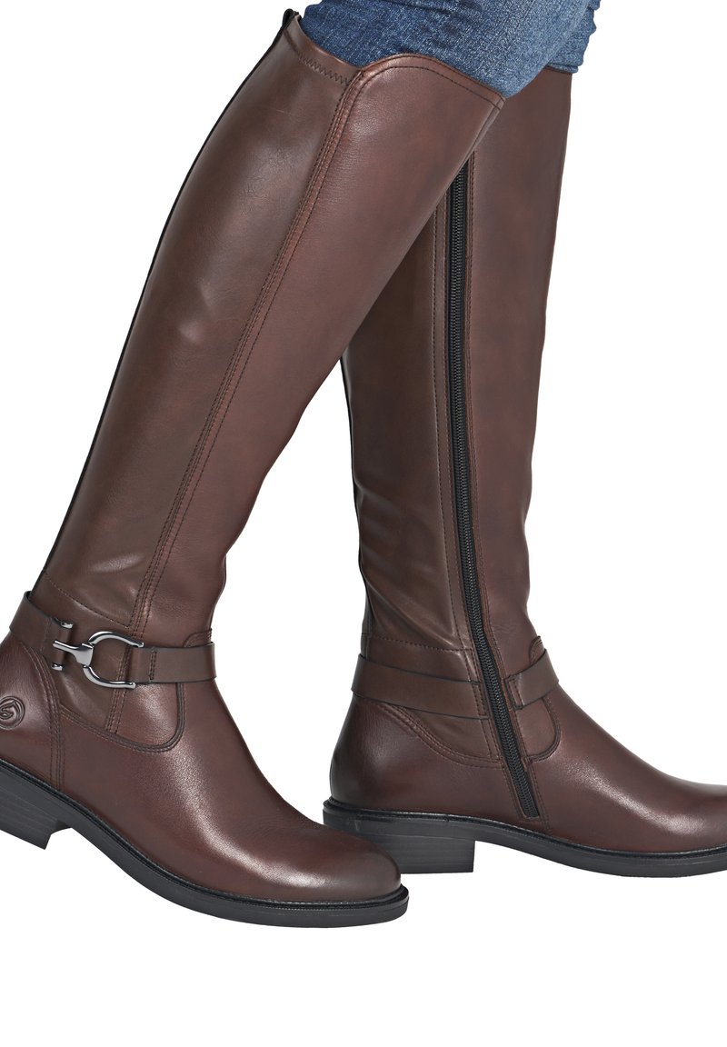 Bottes en cuir marron montant jusqu'au genou avec fermetures éclair latérales noires, sangles décoratives, boucle argentée, portées avec un jean bleu.