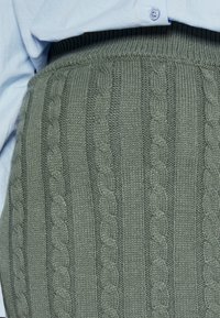Detailní záběr na pletenou látku se zeleným kabelovým vzorem v kombinaci se světle modrou košilí na knoflíky vsunutou dovnitř.
