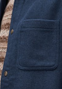 Μπλε μάλλινο μπουφάν με μπροστινή τσέπη, που διαθέτει υφή στην επιφάνεια και στρογγυλεμένες άκρες, συμπληρωμένο με πατιλέτα από ορείχαλκο.