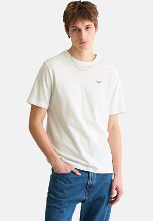 Junger Mann, der ein schlichtes weißes T-Shirt mit kurzen Ärmeln und blaue Jeans trägt, steht mit einer Hand in der Tasche vor einem einfarbigen Hintergrund.