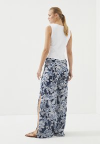 Witte mouwloze geribbelde top gecombineerd met marineblauwe wijde broek met een wit bloemenpatroon en zijsplitten. Bruine sandalen zichtbaar.