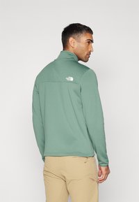 Giacca a pullover verde chiaro con colletto alto, tessuto testurizzato e un piccolo logo bianco sul retro. Abbinata a pantaloni beige.