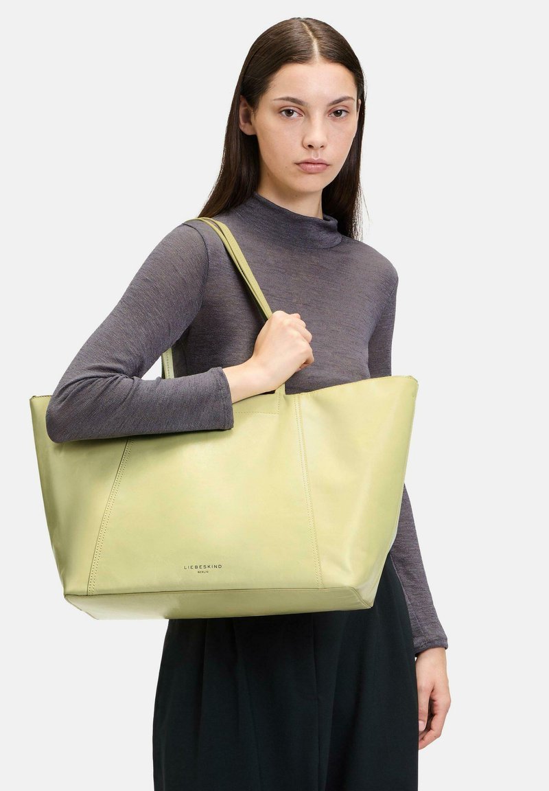 Sac fourre-tout en cuir vert clair avec une forme structurée, des bords supérieurs arrondis et un logo discret à la base. Il est doté de longues bretelles solides.