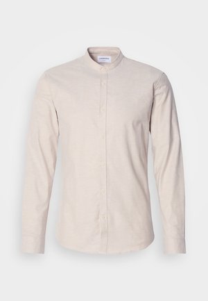 Šviesiai smėlinė ilgos rankovės marškiniai su mandarinų apykakle, rodomi iškyšantiais frontaliame plane ant vientisos baltos fono.