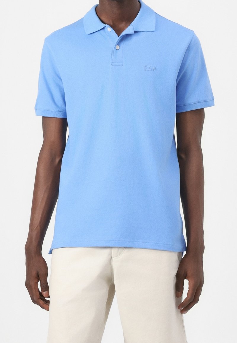 Homme portant un polo bleu clair à manches courtes avec deux boutons et un pantalon beige sur fond uni.