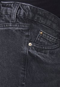 Närbild av svarta denimjeans som visar detaljerad sömnad, bälteögla, metallnitar och en del av framfickan mot en mörk tygbakgrund.