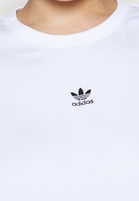 White cotton t-shirt with a ribbed crew neckline, featuring a small black Adidas trefoil logo on the chest. Smooth texture.