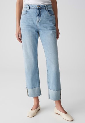 Femme portant un jean bleu clair droit avec de larges revers et des chaussures crème à talons bas, debout devant un fond uni.