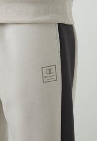 Pantalones deportivos en gris y azul marino oscuro con una textura suave, que presentan un parche con el logotipo de Champion en el muslo izquierdo. Incluyen una cintura elástica.