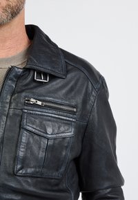 Svart läderjacka med stor krage, dragkedjeförsedd bröstficka, framficka med lock och texturerad yta. Har en spänndetalj på kragen.