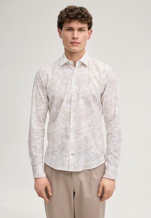 Giovane uomo con capelli ricci che indossa una camicia bianca con fiori e pantaloni beige, in piedi davanti a uno sfondo chiaro e uniforme.