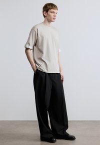 Beiges Baumwoll-T-Shirt mit umgeschlagenen Ärmeln und einem kontrastierenden weißen Streifen, kombiniert mit locker sitzenden schwarzen Hosen und schwarzen Schuhen.