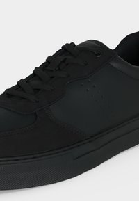 Baskets basses noires avec un design à lacets, panneaux en cuir lisse et perforé, et semelle en caoutchouc texturée sur un fond clair.
