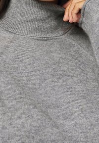 Grauer Rollkragenpullover, gerippte Textur, weiches Material, mit hohem Kragen und Raglanärmeln, Nahaufnahme mit Fokus auf die Stoffdetails.