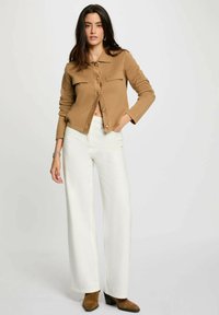 Giacca in maglia cammello con bottoni dorati, due tasche sul petto e collo a trama, abbinata a pantaloni bianchi a vita alta e gamba larga e stivaletti marroni.