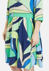 Buntes Kleid mit einem gestuften Design und geometrischen Blattmustern. Die Materialien bestehen aus weichem, leichtem Stoff in Blau-, Grün- und Neutraltönen.