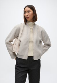 Lys grå ull-blandings bomberjakke med ribbestrikkede mansjetter og nederkant, brukt over en kremfarget ribbestrikket turtleneck, kombinert med mørke bukser og en beige håndveske.