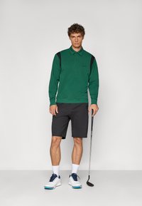 Grønn langermet polo med sorte skulderdetaljer, kombinert med mørke shorts. Modellen holder en golfkølle og har på seg hvite sko med blå detaljer.