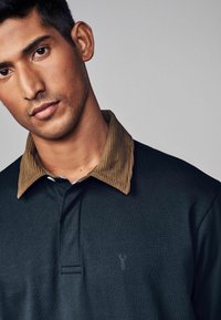 Polo de culoare verde închis cu guler din catifea maro; are un logo subtil pe piept și o deschidere cu plachetă în față. Textură netedă.
