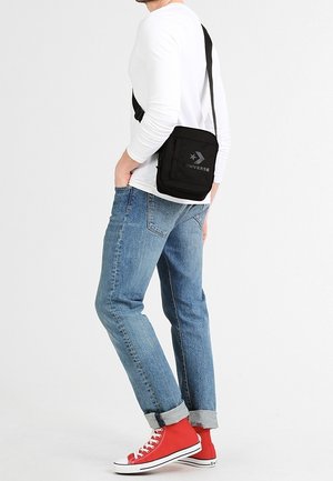 Sac en tissu noir bandoulière avec fermeture éclair et sangle réglable ; arbore un logo d'étoile grise à l'avant. Associé à un jean bleu et des baskets rouges.