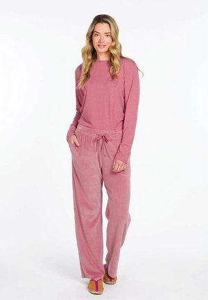 Vrouw staand, gekleed in een roze top met lange mouwen en bijpassende losse roze broek met trekkoord, en beige slippers met paarse bandjes.