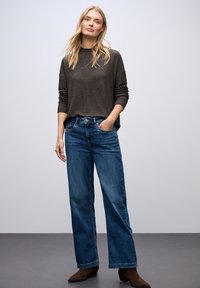 Maglia lunga grigia scura con una vestibilità rilassata; abbinata a jeans blu a gamba larga. Il modello è in piedi con una mano in tasca, indossando stivali alla caviglia marroni.