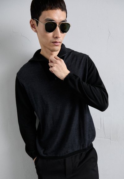 Férfi sötét repülőgép-stílusú napszemüvegben és fekete, hosszú ujjú, zipzáros pulóverben, egyik kezével a zipzárt igazítja egy egyszínű világos fal előtt.