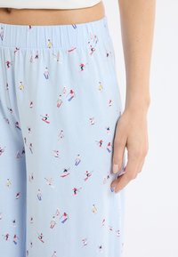 Pantalon de pyjama bleu clair avec une taille élastique, présentant un motif ludique de skieurs et snowboardeurs multicolores sur tout le tissu.