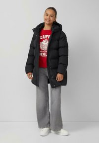 Zwarte pufferjas met een capuchon, met een rits- en drukknoopsluiting. Gecombineerd met een rode graphic sweater en lichtgrijze flared jeans.