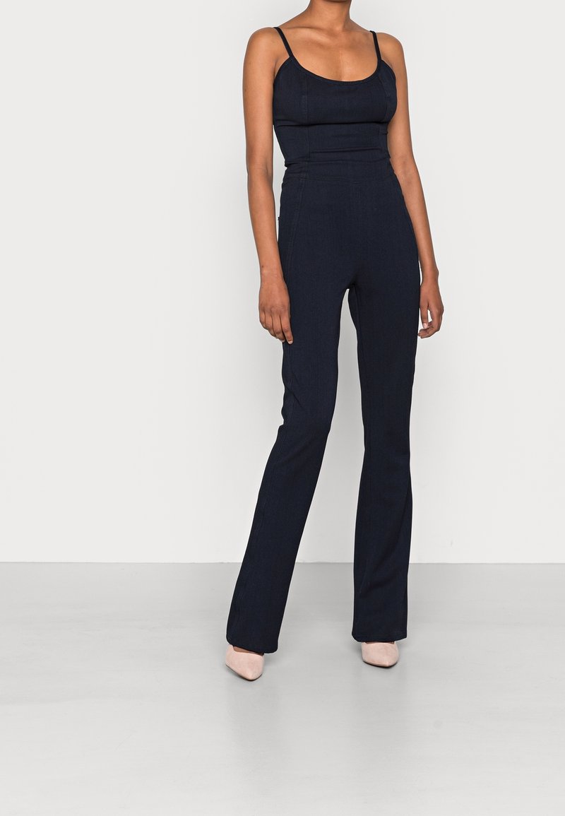 Femme portant une combinaison noire ajustée sans manches avec un pantalon évasé et des chaussures pointues de couleur claire, debout devant un fond clair uni.